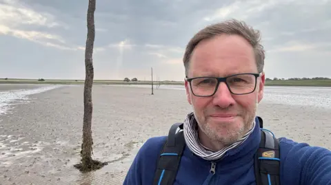Gareth Davies-Jones Gareth Davies-Jones standing on a beach doing a selfie. He has black glasses and short brown hair. Here is a white stubble. He is wearing a blue jacket and a backpack. 