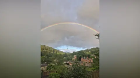 Weather Watchers/Rebecca Blackwell A rainbow in a cloudy sky over a valley with wooded sides and a few houses nestled amidst greenery in the middle distance