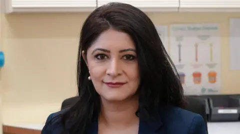SHAUN WHITMORE/BBC Dr Shabina Qayyum wearing a navy blazer and dark blue top is looking at the camera and smiling. Behind her, out of focus, is a GP surgery. 