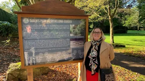 Rachel Lane - wearing beige coat, a scarf, glasses, black top and red skirt, stands in Queen's Park, beside an information board explaining the rare form of cancer