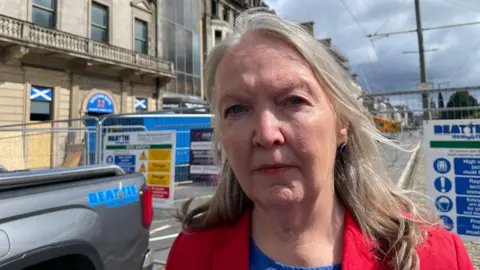 Jane Meagher looking slightly off camera. She is wearing a red jacket over a blue top. She is standing in front of a heras fence and the back of a pick up truck.