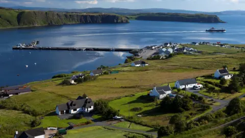 Getty Images White-walled houses and other properties in Uig on Skye. The settlement is on the shores of the sea. There is a Caledonian MacBrayne ferry sailing off from Uig harbour. The land is green and the photo was taken on a fine, bright day.