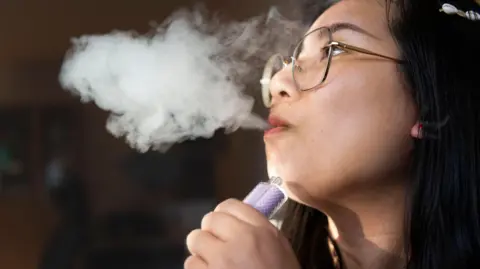 A young person exhaling smoke from a vape, which she is holding in her hand. 