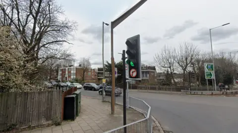 A general view of a pedestrian traffic light crossing on Holders Hill Road, with cars on the road and residential buildings and trees in the background.
