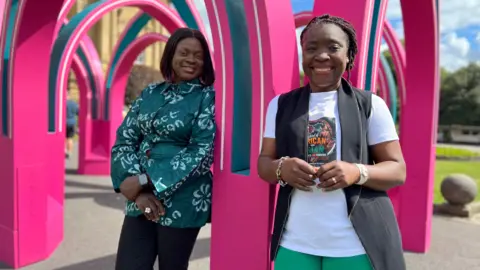 Aisha Iqbal/BBC Two ladies stand smiling underneath a pink arched art structure. They are both black and wear colourful, cultural clothing. One of them wears a T shirt promoting an African and Asian festival.