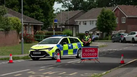 Police officer standing behind cordon on a road with police car behind and homes in the background.