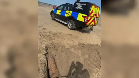 The old navy shell lying in a hole dug by officers on the beach. It is round, long and rusty. On the sand to the right is the shadow of the person taking the photo. There is an emergency service vehicle also in the background parked on the beach with the sea in front of it. In front of it is a red flag and the truck has high-viz livery and the lettering ROYAL NAVY BOMB DISPOSAL.
