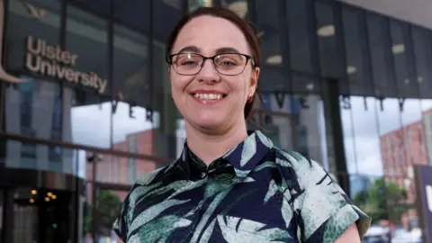 Liam McBurney/PA Wire A woman in a blue shirt with green leaf pattern standing in front of an Ulster University building with glass panels. She has black hair and black glasses.