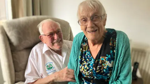 Shariqua Ahmed/BBC Richard, a man in a white shirt, smiling for the camera. While his wife Margaret, in a blue dress and cardigan, sitting on his lap also smiling.