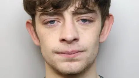 Mugshot of Ronnie. He has stuble around his chin. Floppy brown hair which covers his thick eyebrows. He is standing against a grey backdrop.