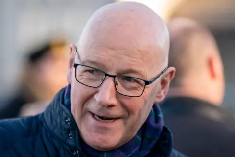 PA Media John Swinney is a bald man with dark glasses wearing a padded navy jacket. Two blurred heads are in the background and Swinney smiles mid-convo while looking off to the side 
