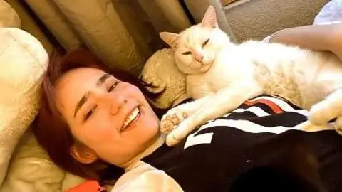 Elise Sebastian lies on a sofa with her cat on her. It is a white short haired cat, Elise is similing and wearing a long sleeved black and white top