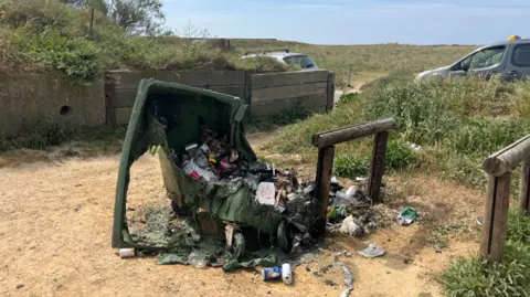 BBC A melted, burnt-out wheelie bin filled with charred rubbish sits on a sandy path in a grassy dune car park, with litter scattered around and cars parked nearby under a clear blue sky.