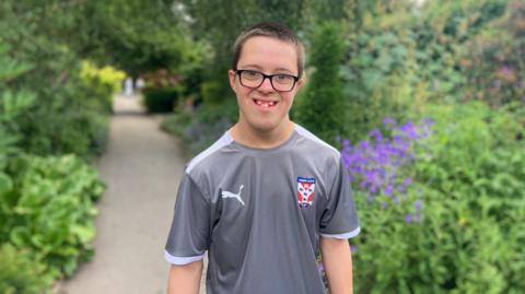 Jack is in a park surrounded by trees and flowers wearing glasses and a grey football shirt with the York City FC logo on his shirt 