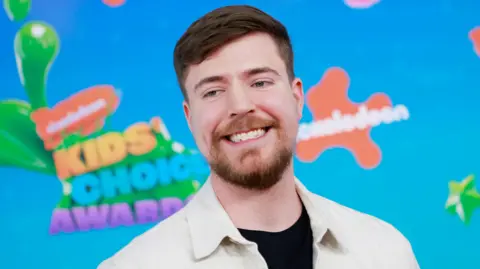 Foto's van Jimmy Donaldson, alias MrBeast, tijdens de Nickelodeon Kids' Choice Awards. MrBeast heeft kort bruin haar en een korte baard. Hij draagt ​​een wit jasje over een zwart overhemd en staat voor een helderblauw bord met het oranje Nickelodeon-logo. 