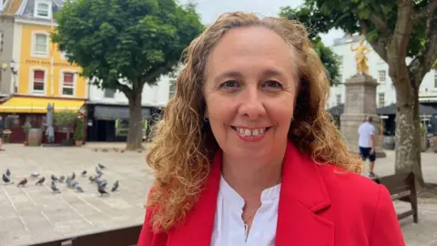 Deputy Inna Gardiner with curly hair wearing a red coat and white shirt, standing outdoors in a town square. In the background, there are trees, buildings, benches, and pigeons on the ground, along with another person walking in the distance.

