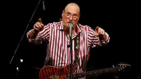Reuters Steve Cropper z brązową gitarą zwisającą z szyi i gestykuluje obiema rękami skierowanymi do przodu. Nosi zapinaną na guziki koszulę w czerwono-białe paski