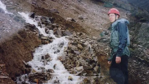 Richard Johnson Professor Richard Johnson, wearing a red helmet, blue coat and trousers, is standing next to large rocks with water cascading down the middle of a crevice in the rock.