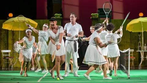 Nilz Böhme  Mr Chaundy is pictured in white trousers and a white polo shirt on stage, surrounded by dancers prancing around him in tennis apparel, brandishing racquets