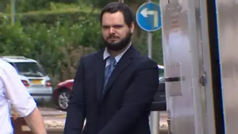 BBC Kyle Enos arriving at court wearing navy suit and light blue tie and shirt. He has a dark beard and dark short hair and is handcuffed and walking behind a prison officer in white short-sleeved shirt.