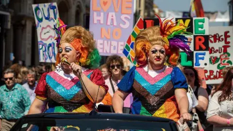 Tekaani Media Colourful drag queens in a convertible car lead the crowds through the streets at Pride Canterbury in 2023