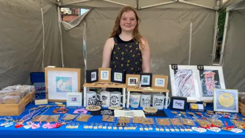 Cumberland Council Teenager in flower dress stands behind stall filled with mugs, artwork and jewelry
