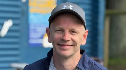 A smiling man in a blue cap in front of a skate park coffee shop.