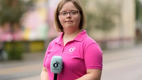 TV BRA A young woman with Down syndrome stands on the street, holding a microphone. She wore a bright pink polo shirt. Her hair is medium brown and doesn't reach her shoulders. She wore glasses and smiled. 