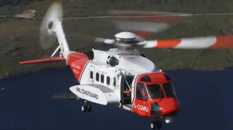 A red and white HM Coastguard helicopter in flight over some coastline.