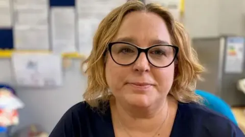 A middle aged woman with shoulder length wavy blonde hair and dark rimmed glasses smiles at the camera