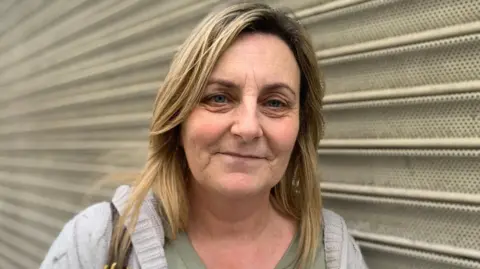 A woman with long blonde hair looks into the camera. She is leaning up against grey metal shutters. She wears a grey jumper and green t-shirt.