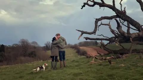 Courtesy of Prince Harry and Meghan, The Duke and Duchess of Sussex A handout photo shows the Duke and Duchess of Sussex with their late dog, Guy. The two are standing in a field in the middle distance with their backs to the camera. The duke's left arm is round the duchess's shoulders and the two are kissing. Guy is on a lead held by the duchess and is standing next to them. 