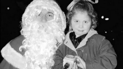 Cambs Times/Newsquest A black and white picture of a five-year-old girl standing next to Santa, the girl is holding a wired box controller to switch the lights on for the Christmas display. She has dark hair, with a ribbon tied in the top, she is wearing a fur lined jacket over a dark top. Santa's face is almost entirely covered by a white beard and hair.