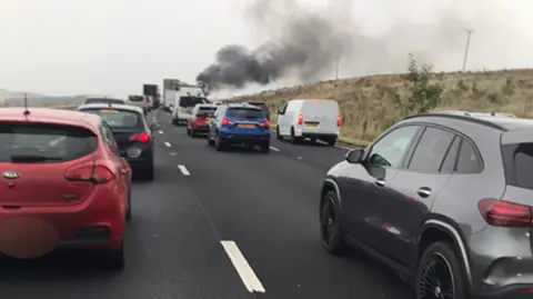 Chris Isles The M6 lined with cars across three lanes. Black smoke can be seen rising in the distance. 