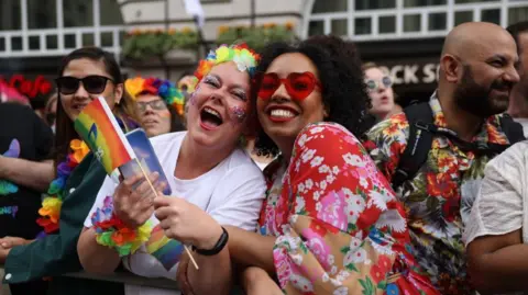 A picture focused on two attendees of a previous Pride in London event. On the left, a woman with glitter on her face and with flowery decorations around her head and arms laughs at the camera. On the right, another woman smiles at the camera with red-heart sunglasses on and a red flowery dress.