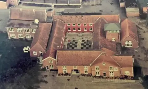 Shirley Oaks Survivors Association Aerial view of Red House at Buxton, Norfolk, featuring a quadrangle of red brick buildings and ancillary buildings. There is a chequerboard pattern in the centre of the quadrangle, and greenery to the lower left of the image, and hard standing on all other elevations.