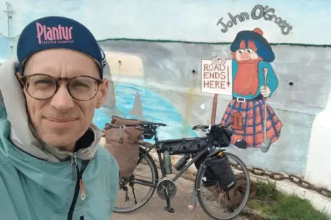 Christoph Held Christoph Held is wearing a navy "Plantur" cap and a light blue coat. His bike is behind him leaning against a wall with a painting of a ginger haired man wearing a kilt holding a sign saying "Road ends here". Above the painting are the words "John O'GRoats".