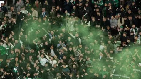 Hibs fans in the stands during their game vs Hearts. Green smoke, released by pyro and smoke devices, is going across the fans.