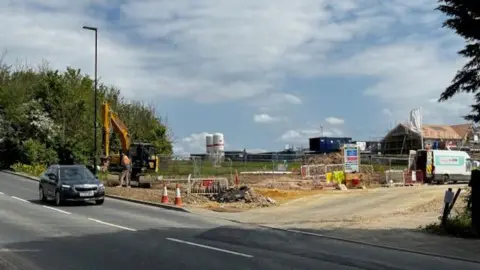 Picture shows a building site, with a road in the foreground.