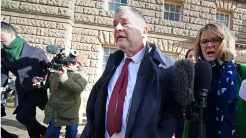 PA Media Mike Amesbury, wearing a navy blue coat over a white shirt and burgundy tie, walks away from court flanked by camera operators and journalists.