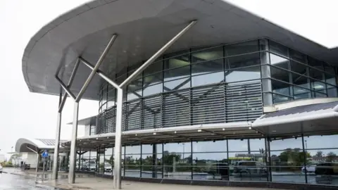 The exterior of Guernsey Airport's main terminal building which consists of dozens of windows and a large white roof which overhangs the main section of the building and is being held up by three silver y-shaped posts.