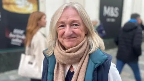 Janet Wild in Strand Street smiling at the camera. She has blonde hair and is wearing a blue body warmer and beige scarf.