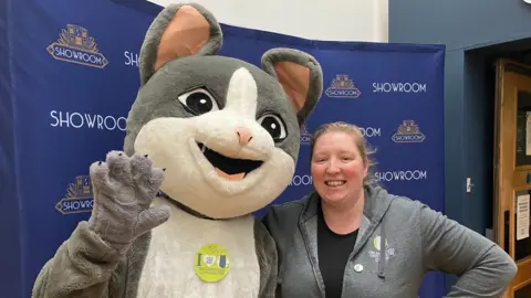 Someone dressed as a large grey and white cat and a woman with tied back blonde hair wearing a black top and grey hoodie. They stand in front of a blue boar that reads: Showroom.