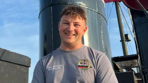 Chris Bannister A smiling man, wearing a grey t-shirt with a logo of a vessel, is standing on a boat.