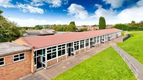 A long single-storey building with lots of windows and a red roof, with a footpath and a grassy area running alongside