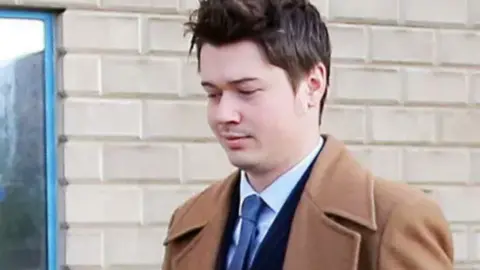 SWNS A man with dark hair, a dark suit, blue tie and brown coat standing in front of a white brick wall
