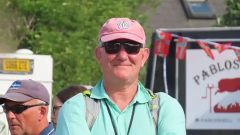 Lt Gen Richard Cripwell - A white man with a red baseball cap on, alongside black sunglasses. He is wearing a turquoise shirt and a grey back pack and smiling at the camera.