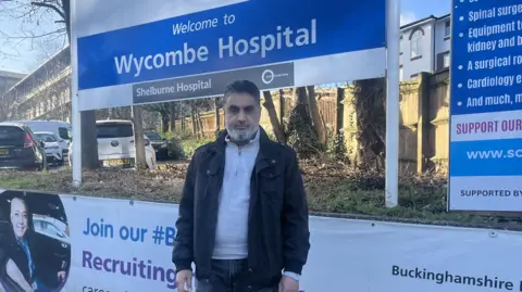 Khalil Ahmed Khalil Ahmed wearing a dark jacket stands in front of a sign reading “Welcome to Wycombe Hospital” on a sunny day, with hospital buildings and parked cars behind him.