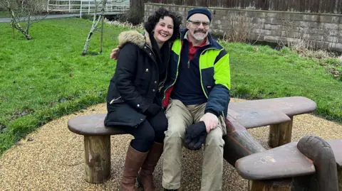 Two people, a man and a woman, sit on a wooden carved bench in the shape of a World War Two Barracuda plane