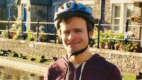 Family handout Max McGhee wearing a hoody and a bike helmet. He is stood next to a river and there are stone houses behind him. 